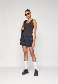 Sort tanktop, marineblå shorts med to sidelommer, gul snor, hvide ankelstrømper og sorte træningssko. Brune solbriller.