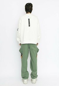 Sudadera blanca de gran tamaño con etiqueta negra, combinada con pantalones cargo verdes que tienen bolsillos laterales y acentos bordados, llevados con zapatillas blancas de suela gruesa.