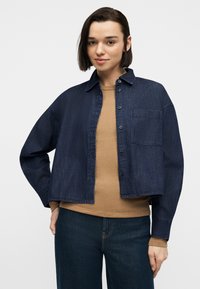 Γυναίκα με κοντά σκουρόχρωμα μαλλιά φοράει ένα κοντομάνικο σκούρο τζιν μπουφάν πάνω από ένα μπεζ ριμπ πλεκτό πουλόβερ και σκούρο τζιν, ποζάροντας με το ένα χέρι στη μέση.