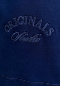 Námorně modrý svetr s texturovanou výšivkou se slovy "ORIGINALS Studio" v kurzívě, zdůrazňující design značky.