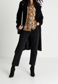 Cappotto lungo nero sopra una camicia a fantasia con stampa pitone in toni caldi. Abbinato a jeans neri e stivali alla caviglia, creando un look a strati.