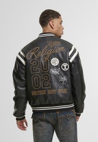 Juodas odinis sportinis švarkas su baltais juostelėmis ant rankovių. Papuoštas auksinėmis siuvinėjimo detalėmis ir keliais pleistrais, įskaitant "True Religion" ir numeruotus elementus.