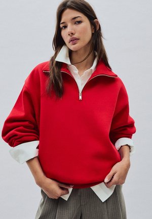 Jonge vrouw die een rode trui met halve rits draagt over een wit overhemd met opgerolde mouwen en een grijze broek, staand tegen een effen achtergrond.