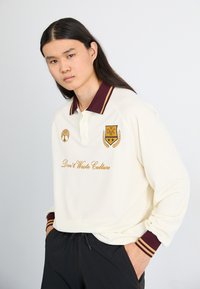 Polo crème avec col bordeaux, orné d'une broderie dorée, texte "Ne gaspillez pas la culture" et logo. Manches longues avec poignets rayés.