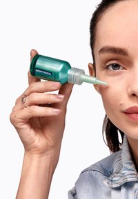 Bouteille turquoise avec un embout applicateur pointu étiqueté « RÉVÉLATION ÉCLAT ». L'utilisateur applique le produit sous l'œil ; la peau est claire avec une texture naturelle.