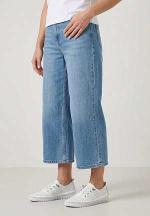Person iført lyseblå vide korte jeans med hvide perforerede sneakers, der står på en ensartet lys overflade.