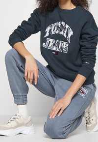 Sudadera oscura con el logo "TOMMY JEANS", combinada con pantalones de chándal gris claro. El calzado consiste en zapatillas crema con toques de colores múltiples.
