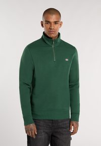 Groene sweatshirt met kwart-rits, gemaakt van dik materiaal, met een geribbelde kraag en boorden. Bevat een klein logo op de borst.