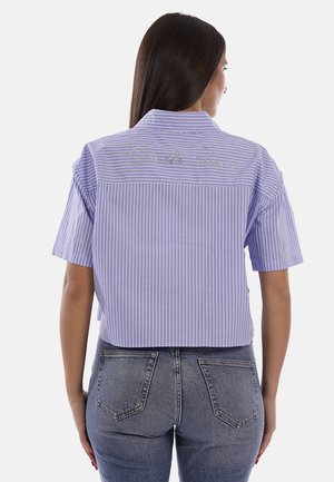 Donna con capelli lunghi e scuri indossa una camicia a maniche corte a righe azzurro chiaro e bianco con testo in strass sulla schiena, abbinata a jeans di denim blu.