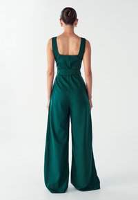 Mujer vista desde atrás, vistiendo un mono verde sin mangas y de pierna ancha, con un escote cuadrado en la espalda y aretes de aro dorados.