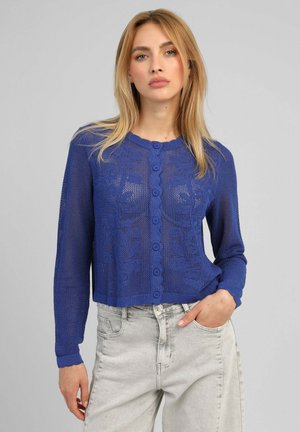 Femme blonde portant un cardigan bleu à motifs boutonné et un jean gris clair taille haute, debout avec une main dans la poche.