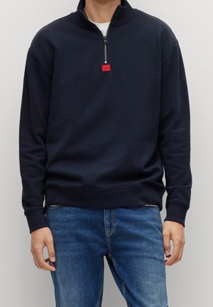 Mörkblå zip-up sweatshirt med hög krage, med en röd etikett på dragkedjan, tillverkad av mjukt tyg. Kombinerad med ljusblå jeans.