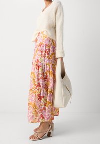 Pull en tricot crème avec un col en V, associé à une jupe maxi à motifs floraux roses. Des sandales à lanières beiges et un sac à main blanc complètent le look.