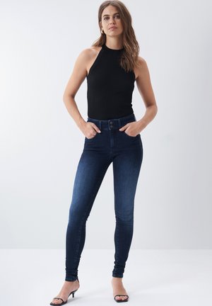 Vaqueros skinnies de tiro alto en azul oscuro, hechos de mezclilla, con cierre de botón y dos bolsillos frontales, combinados con una blusa negra ajustada sin mangas.