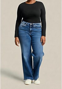 Femme portant un haut noir à manches longues, un jean bleu taille haute à jambes larges et des baskets blanches, debout devant un fond uni.