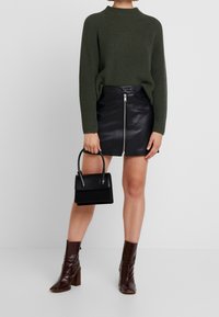 Person trägt einen dunkelgrünen Strickpullover und einen schwarzen Lederrock mit Frontreißverschluss, hält eine kleine schwarze Handtasche und kombiniert dazu braune Stiefeletten.