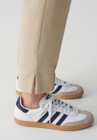 Witte Adidas Samba sneakers met blauwe strepen en grijze suède accenten, gecombineerd met beige broeken met een split aan de zoom.