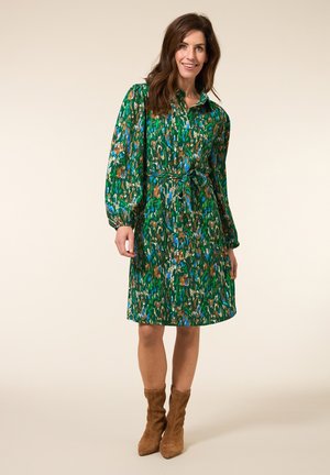 Groene jurk met abstracte blauwe en bruine patronen, lange mouwen, knoopsluiting en tailleband. Draagt met tan enkelboots.