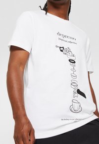 Valkoinen puuvillapaita, jossa on mustia graafisia kuvastoja kahvinkeiton osista ja teksti, joka sisältää humoristisia määritelmiä kahvista.