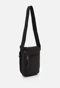 Bolso de hombro negro hecho de tela, con un bolsillo frontal con cremallera, correa ajustable y un acento de cadena en el tirador de la cremallera.