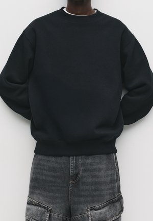 Person iført simpel sort crewneck-sweatshirt og falmede sorte denimshorts mod en hvid baggrund.