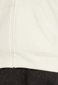 Vit bomullshoodie med framficka, synliga sömmar och ribbad kant, kombinerad med mörka, texturerade byxor.