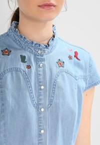 Light blue denim shirt with a ruffled collar, featuring embroidered stars and other motifs, and button closure down the front.