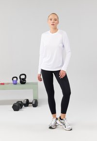 Bela športna majica z dolgimi rokavi, črne legice in srebrni tekaški čevlji. Vadbeni pripomočki vključujejo uteži in kettlebelle na zeleni mizi.