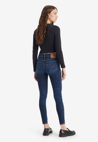 Mujer con cabello largo y ondulado, vestida con jeans skinny azul oscuro y una blusa negra ajustada de manga larga, de pie y de perfil contra un fondo blanco.