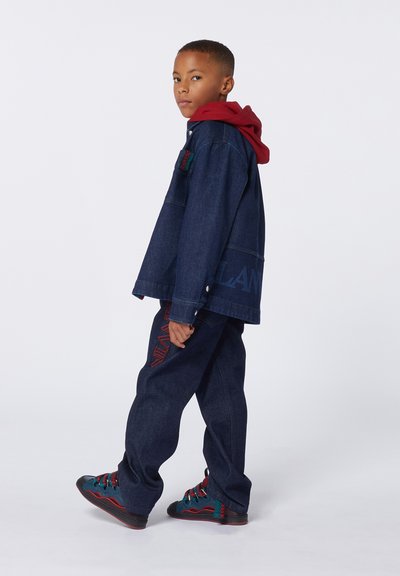 Giacca di jeans con una felpa con cappuccio rossa sotto, abbinata a pantaloni in denim dal taglio largo con ricami rossi e scarpe sportive colorate.