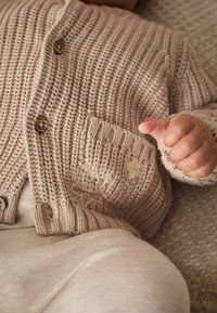 Beige Strickjacke mit vertikalen Streifen, ausgestattet mit Holzknöpfen und einer kleinen Tasche; leichtes Gewebe darunter. Die Hand eines Babys ist sichtbar.