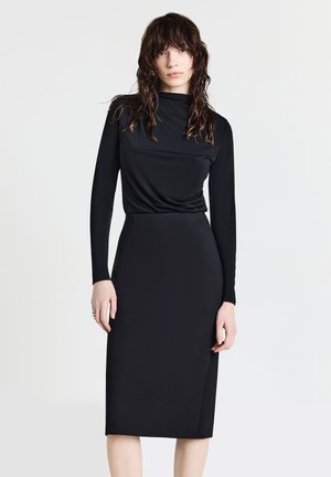 Femme avec les cheveux mouillés portant un haut noir à manches longues rentré dans une jupe noire ajustée longueur genoux sur un fond blanc uni.