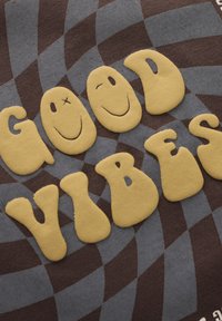 Textiel met "GOOD VIBES" in gele, speelse letters. De achtergrond heeft afwisselend donkerbruine en grijze ruitenpatronen. Zachte textuur.