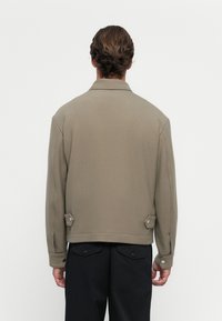Veste kaki avec un col structuré, des manches longues et des accents de boutons aux poignets et sur les côtés, fabriquée à partir d'un tissu lisse.