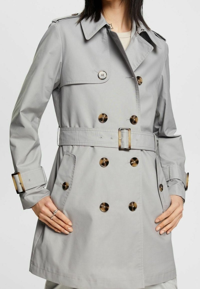 Femme portant un trench-coat gris clair à double boutonnage avec des boutons en écaille et une ceinture à la taille, les mains posées sur les hanches.