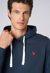 Felpa blu navy in cotone, con finitura texturizzata, dotata di un cordino bianco e di un logo ricamato rosso sul petto.