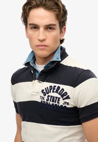Poloshirt met marineblauwe en crèmekleurige horizontale strepen, denim kraag en geborduurde tekst "SUPERDRY TOK. STATE JPN. ATHLETICS" op de borst.