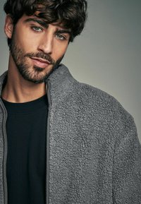 Giacca in fleece grigia con superficie testurizzata, dotata di un alto colletto e chiusura con zip, indossata sopra una maglietta nera a girocollo.