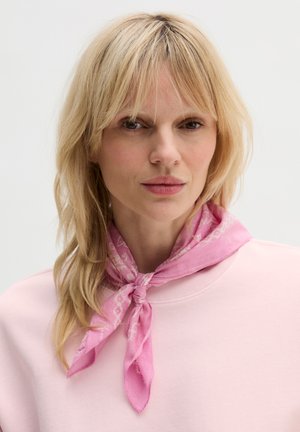 Blonde woman with light makeup wears a pink patterned scarf tied around her neck and a pale pink top against a white background.