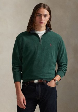 Jeune homme aux cheveux mi-longs portant un pull vert foncé à fermeture éclair quart par-dessus une chemise blanche et un jean foncé avec une ceinture marron et une montre.