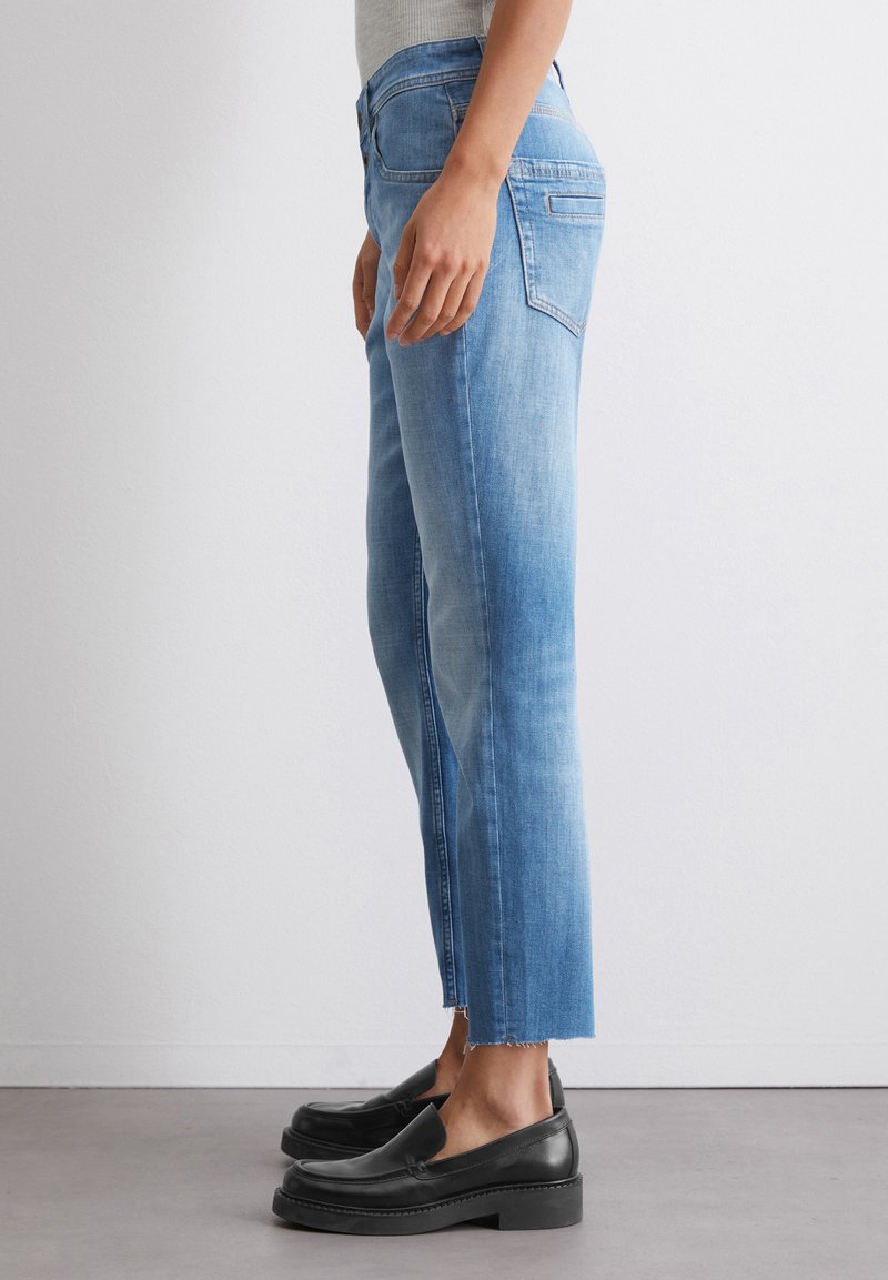 Helle blaue Denim-Jeans mit geradem Schnitt und fransigem Saum. Sie besitzen typische Gesäßtaschen und eine leichte Ausbleichung, kombiniert mit schwarzen Slippern.