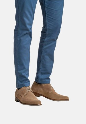 Zapatos de vestir marrones con textura, superficie de malla y cordones, combinados con pantalones ajustados azules, mostrados desde las rodillas hacia abajo sobre un fondo blanco.