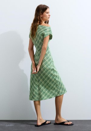 Frau mit langen Haaren, die ein grünes kariertes schulterfreies Midikleid und schwarze Sandalen trägt, steht vor einer hellgrauen Wand.