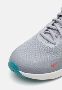 Zapatilla deportiva gris claro con parte superior de malla texturizada, logotipo de Nike en naranja brillante y suela de goma turquesa para tracción.