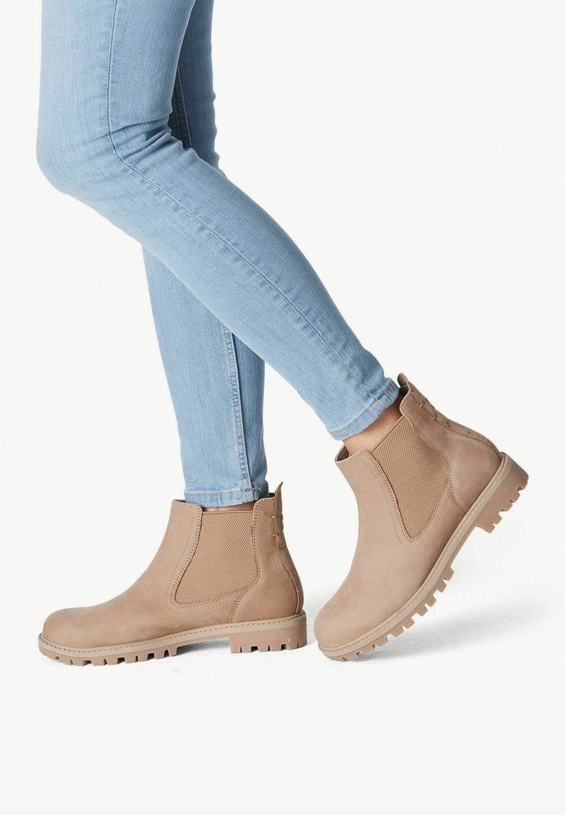 Botas de tobillo de ante beige con paneles laterales elásticos y suelas de goma, combinadas con jeans ajustados de un azul claro. El diseño de las botas presenta detalles sutiles.