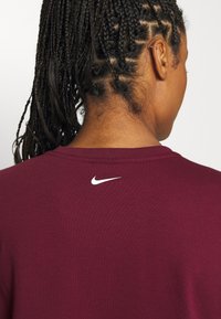 Osoba s copy nosí vínovou košili Nike, přičemž je vidět zadní část výstřihu a malá bílá značka Nike pod límcem.