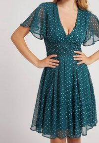 Turkoosi polvenpituisen mekon, jossa on valkoisia pilkkuja, V-kaula-aukko ja liehuvat hihat, päällä henkilö, jonka kädet ovat lanteilla, tasaisessa taustassa.