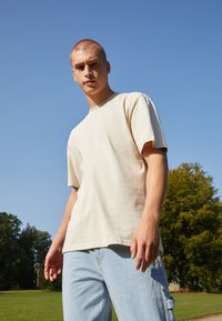 Beige cotton t-shirt with short sleeves, round neckline, and subtle stitching detail; worn with light blue denim. Green grass and trees in background.