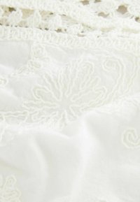 White embroidered fabric featuring intricate floral and spiral patterns, with a textured, layered design and lace accents along the edge.