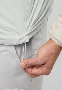 Giacca grigia chiara con cordino elastico all'orlo, che mostra una mano che regola il cordino. Texture del tessuto morbido e liscio.